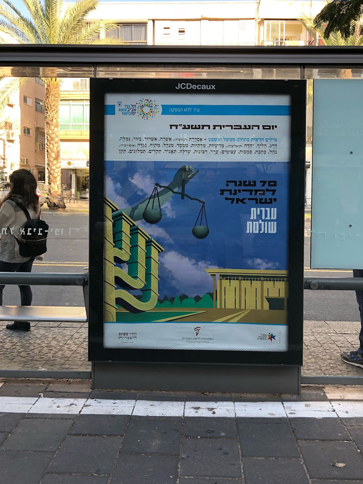 צילום כרזה ליום העברית תשע"ח איור של הכנסת, מנורה, ויד מחזיקה מאזניים וכיתוב "עברית שולטת"