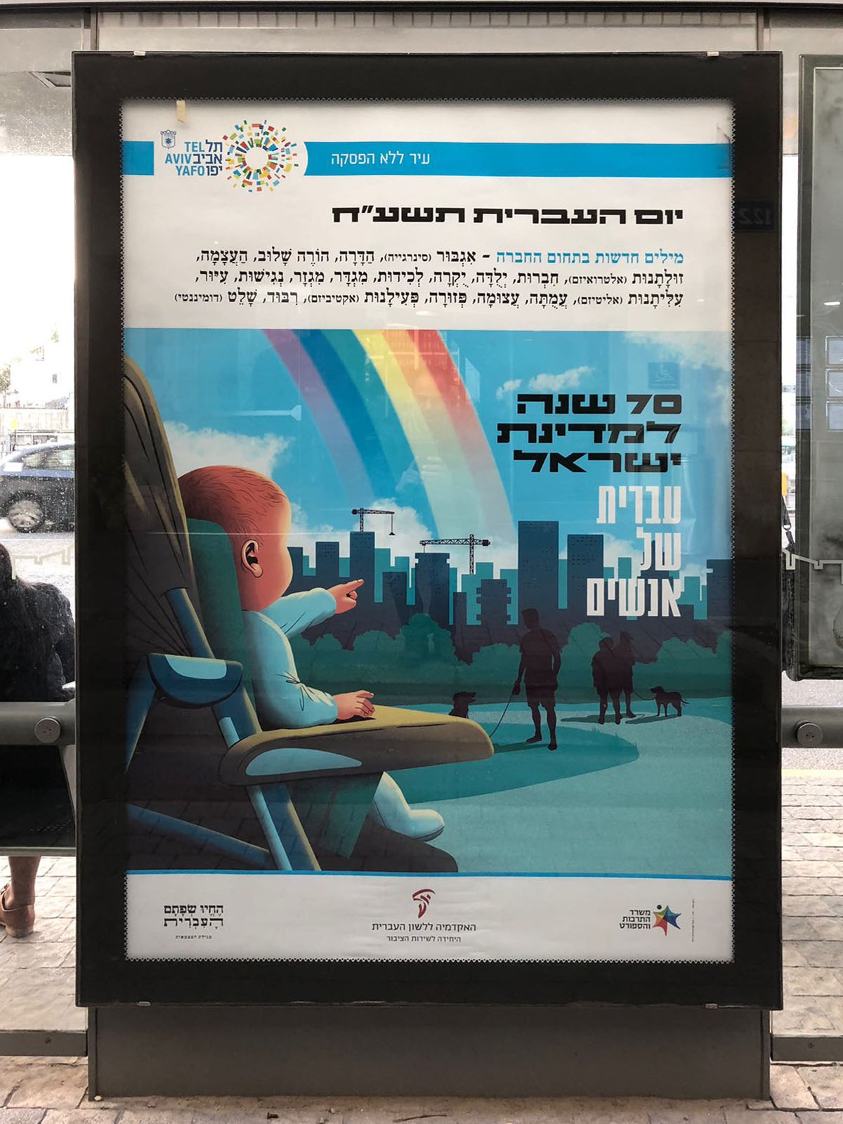 צילום כרזה ליום העברית תשע"ח איור של תינוק בכיסא ילדים מצביע על עיר וכלבים בפארק וכיתוב "עברית של אנשים"